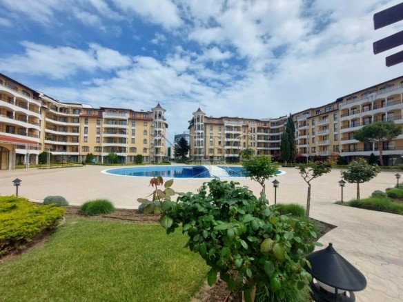 Courtyard of a resort with a circular blue pool, beige apartment buildings, and green landscaping under a partly cloudy sky