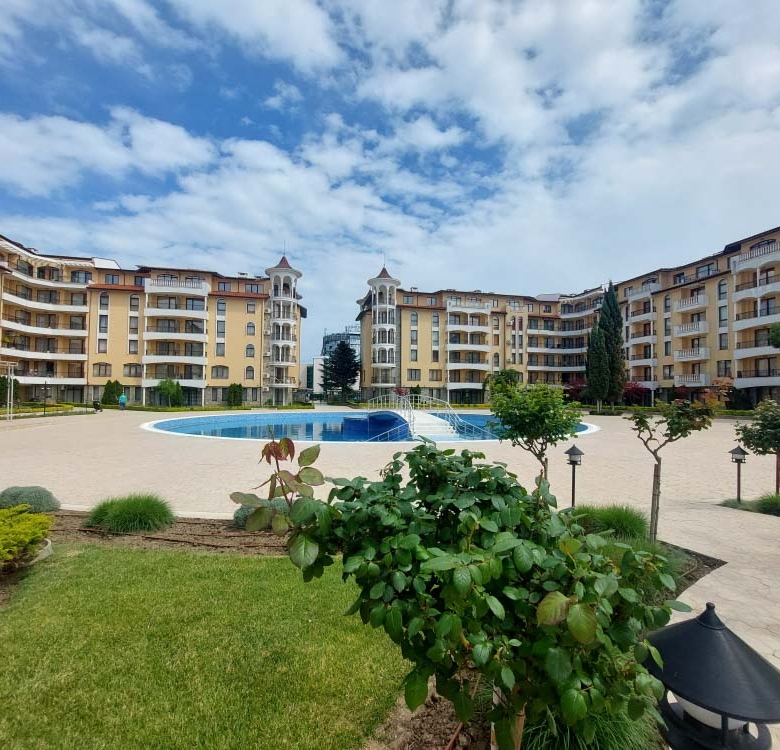 Courtyard of a resort with a circular blue pool, beige apartment buildings, and green landscaping under a partly cloudy sky