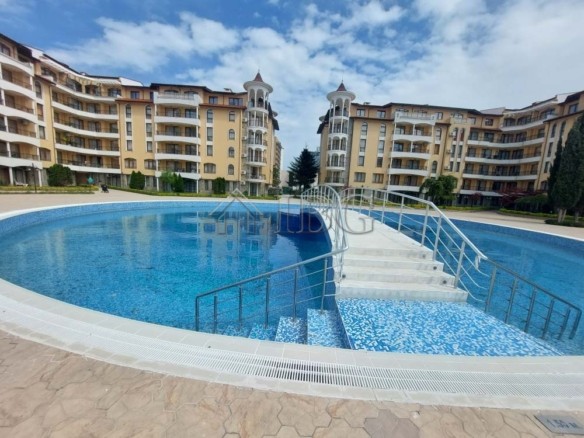 Circular blue pool with a tiled entry bridge and metal railings in front of beige apartment buildings under a partly cloudy sky.