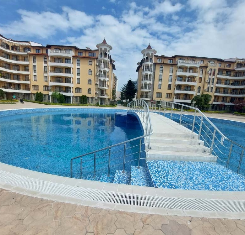 Circular blue pool with a tiled entry bridge and metal railings in front of beige apartment buildings under a partly cloudy sky.