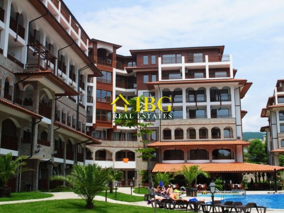 Resort complex with white walls and dark wooden balconies, arches, and a pool area with sun loungers and palm trees in the foreground.