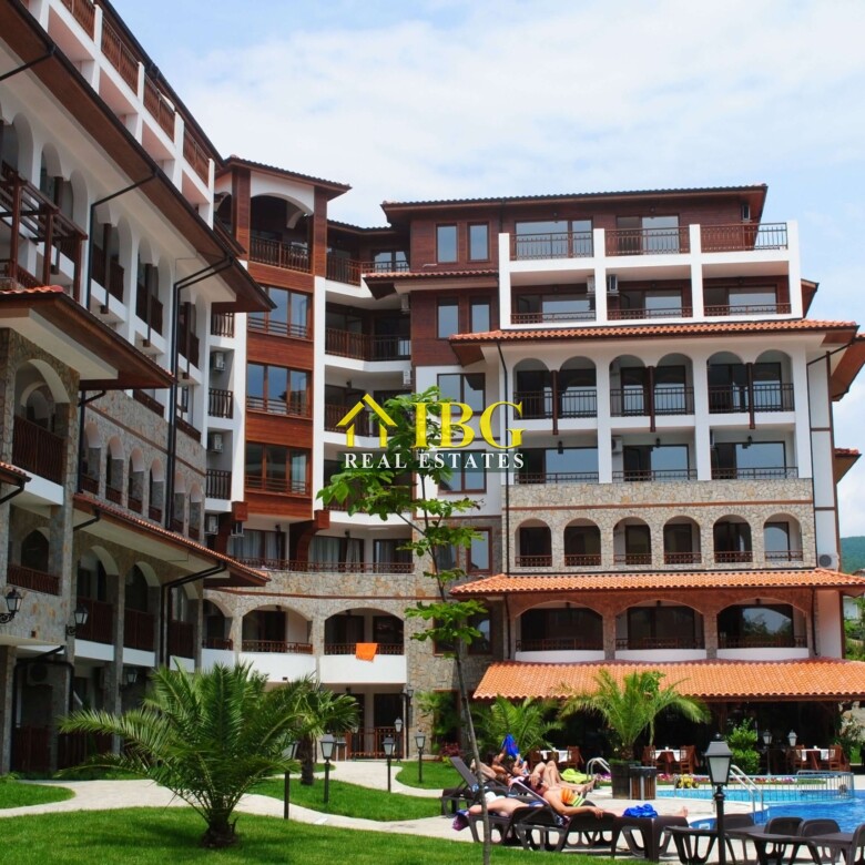 Resort complex with white walls and dark wooden balconies, arches, and a pool area with sun loungers and palm trees in the foreground.