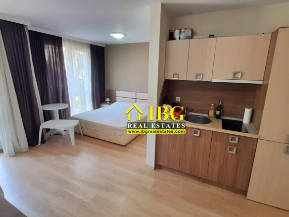 Compact studio room with a kitchenette on the right, a bed against the back wall, and balcony doors with white and dark curtains