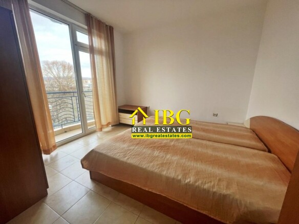 Bright bedroom with a large wooden bed, a small bedside table, wardrobe, and a balcony door with sheer beige curtains.