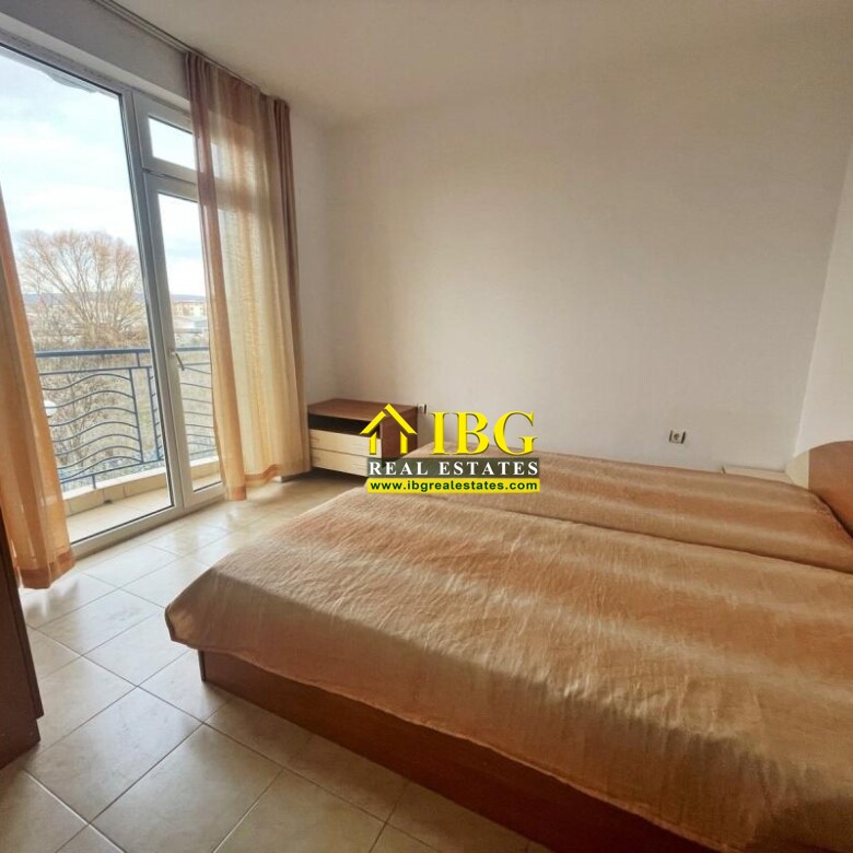 Bright bedroom with a large wooden bed, a small bedside table, wardrobe, and a balcony door with sheer beige curtains.