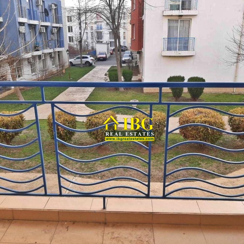 Blue metal balcony railing with wavy horizontal bars, overlooking a courtyard with trees and walkways.