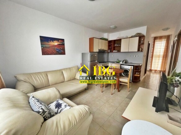 Open-plan living room and kitchen: beige leather sofas, round wooden dining table with chairs, fridge and washing machine, TV, and sunset beach artwork on the wall.
