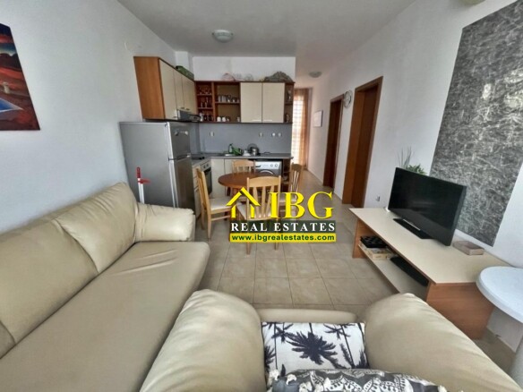 Open-plan living room with a beige L-shaped sofa, TV stand, dining table, and a compact kitchen in the background.