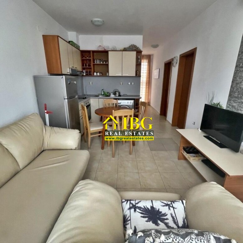 Open-plan living room with a beige L-shaped sofa, TV stand, dining table, and a compact kitchen in the background.