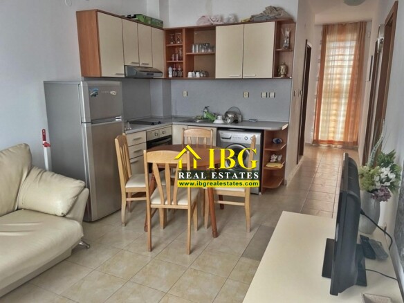 Open-plan kitchenette with a wooden dining table and four chairs, a silver fridge, and a beige sofa nearby.