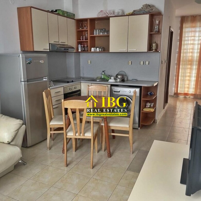Open-plan kitchenette with a wooden dining table and four chairs, a silver fridge, and a beige sofa nearby.