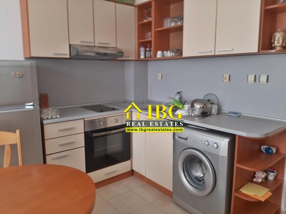 Compact L-shaped kitchen with beige cabinets, stainless steel fridge, built-in oven and gas hob, washing machine, and a round wooden table in the foreground.