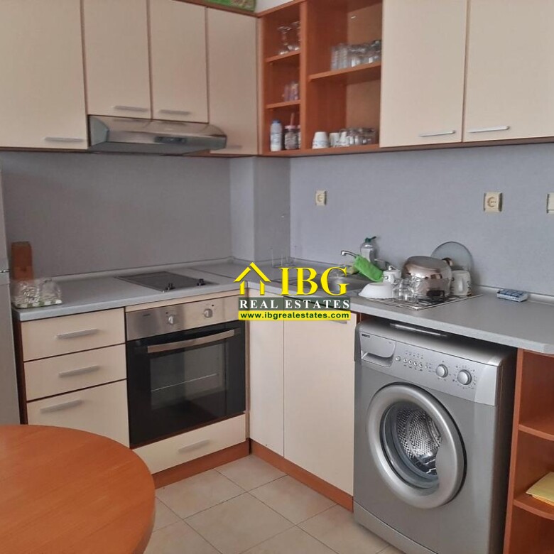 Compact L-shaped kitchen with beige cabinets, stainless steel fridge, built-in oven and gas hob, washing machine, and a round wooden table in the foreground.