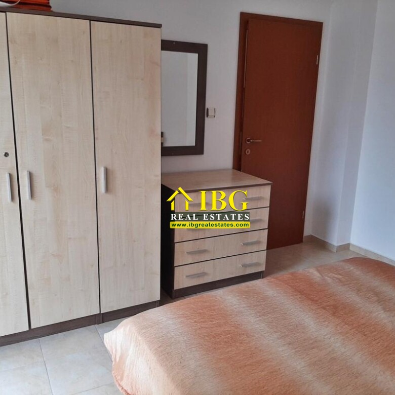 Bedroom with light wood wardrobe, matching chest of drawers, wall mirror, brown door, and an orange blanket-covered bed on tiled floor.