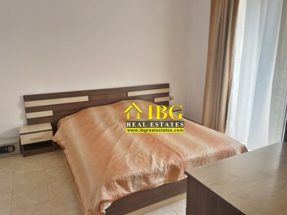 Double bed in a minimalist bedroom with a peach blanket, two matching nightstands, and a wooden headboard by a large window with beige curtains.