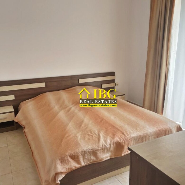 Double bed in a minimalist bedroom with a peach blanket, two matching nightstands, and a wooden headboard by a large window with beige curtains.