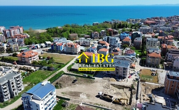 Aerial view of a coastal town with colorful apartment buildings, a construction site, green spaces, and the blue sea on the horizon.